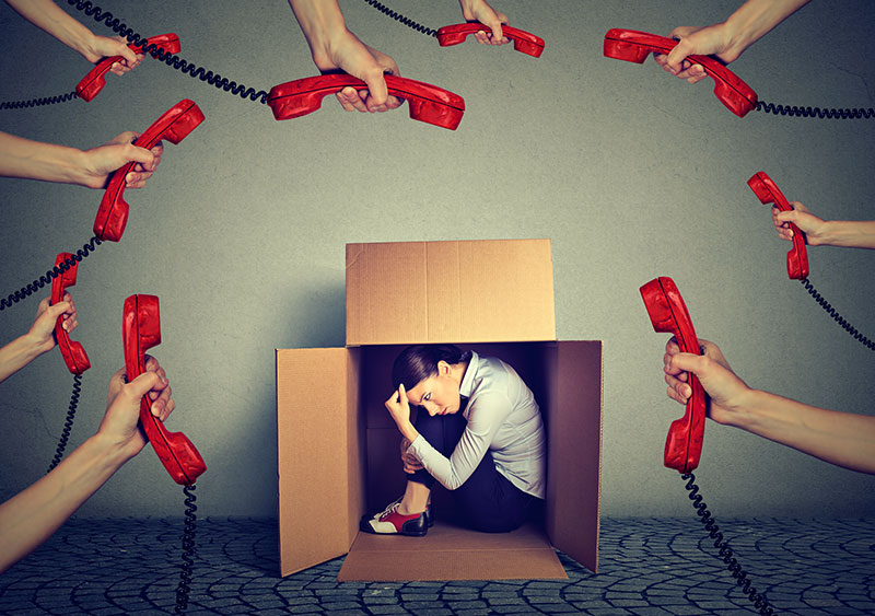Woman sitting inside a cardboard box with red phones ringing all around feeling burned out
