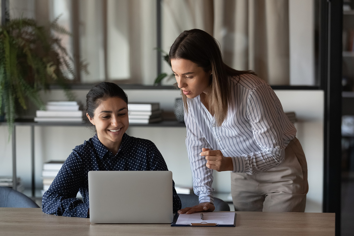 Happy Indian office employee showing job result to boss. Female coworkers discussing project at laptop, talking, laughing. Mentor training intern, supervising work of trainee, explaining task Happy Indian office employee showing job result to boss. Female coworkers discussing project at laptop, talking, laughing. Mentor training intern, supervising work of trainee, explaining task